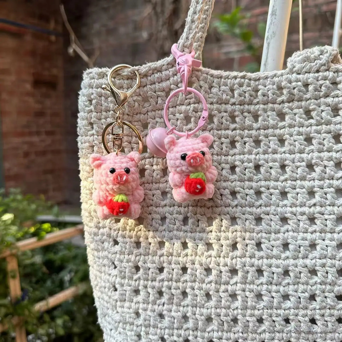 A handmade crochet mini pig keychain with pink ears and a red nose hanging from a gold-colored ring on a beige woven bag.