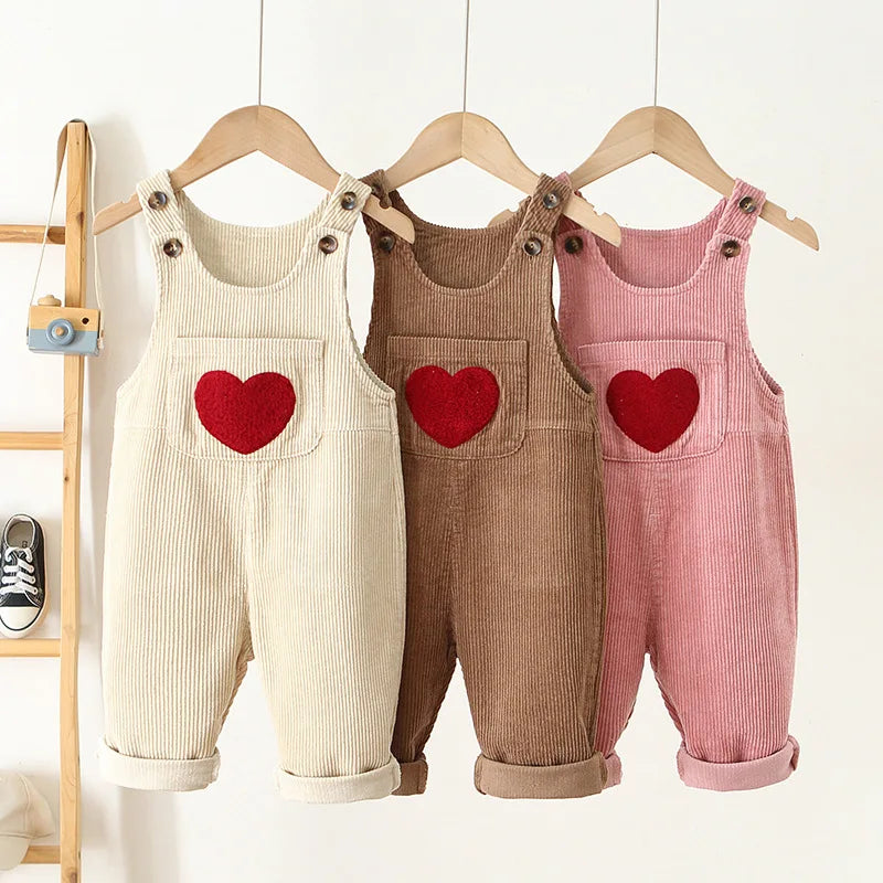 Three pairs of toddler corduroy overalls in cream, brown, and pink hanging on a wooden rack.