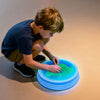 A young boy is interacting with an interactive LED balance stone mat, which displays a green and blue liquid surface.