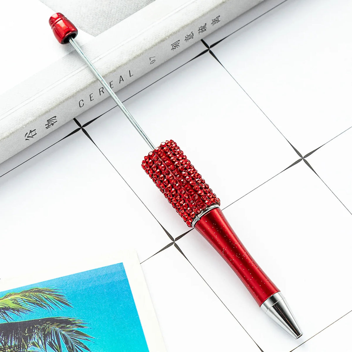 A red beaded ballpoint pen is shown resting on a white surface next to a notebook.