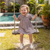 A toddler girl in a plaid A-line dress with a Peter Pan collar is sitting on a swing outdoors.