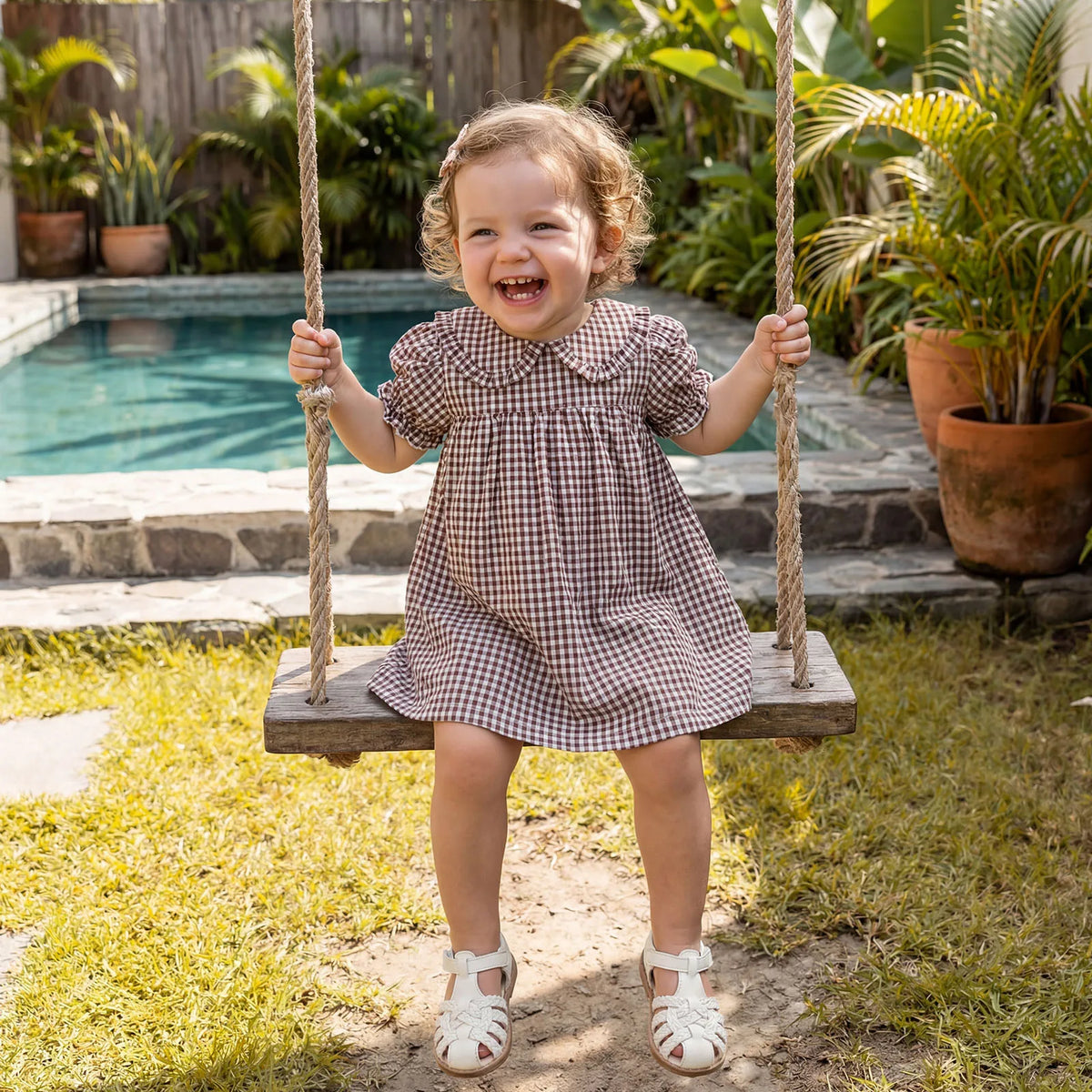 A toddler girl in a plaid A-line dress with a Peter Pan collar is sitting on a swing outdoors.