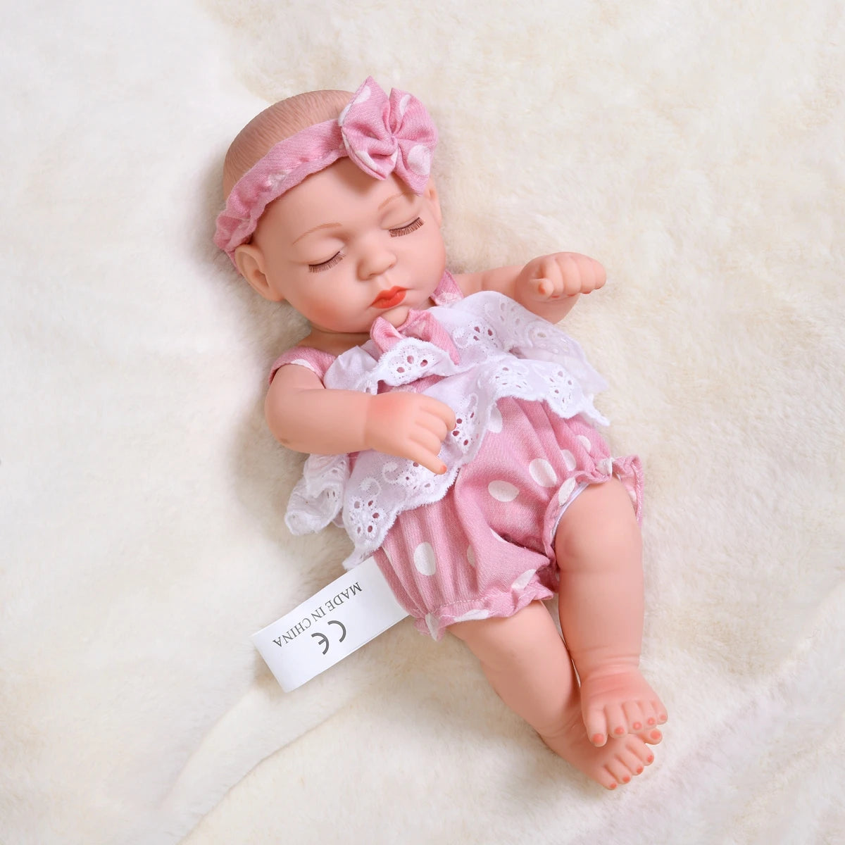 A lifelike soft vinyl reborn doll with a pink outfit and flower headband lying on a white fluffy surface.