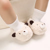 A pair of soft, cream-colored cartoon baby booties with dark accents, shown on a person's foot wearing white socks.
