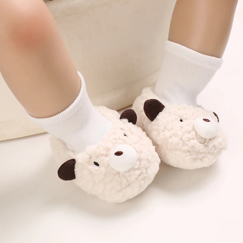 A pair of soft, cream-colored cartoon baby booties with dark accents, shown on a person's foot wearing white socks.