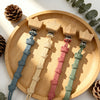 A set of baby pacifier clips in various pastel colors, including blue, beige, pink, and green, displayed on a wooden plate.