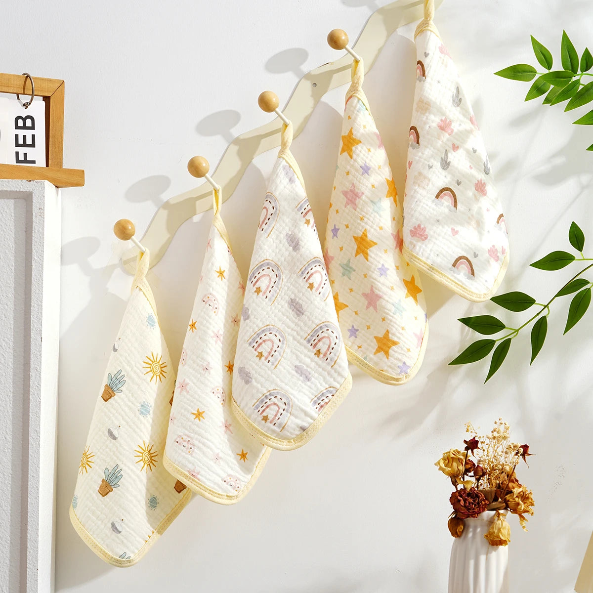 A set of five white and yellow patterned soft cotton muslin baby washcloths hanging on a light wooden rack.