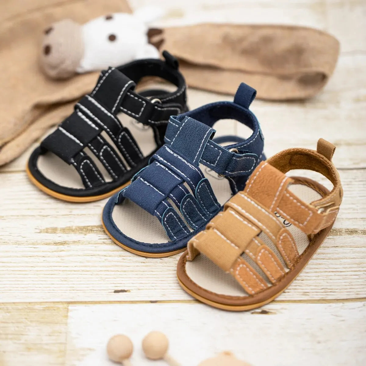 A set of soft non-slip baby sandals for first walkers in various earthy tones including black, blue, and tan.
