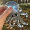 A hand holds a clear, stretchable octopus stress ball with pink and green star decorations outdoors.