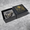 A black box containing a revolver-shaped metal dice set for RPG games with gold accents.