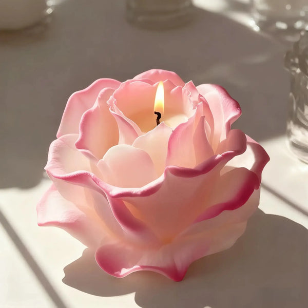 A pink candle in a silicone petal mold sits on a white surface, casting shadows.