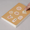 A hand places a finger on the cover of a kraft paper DIY notebook featuring various citrus and floral designs.