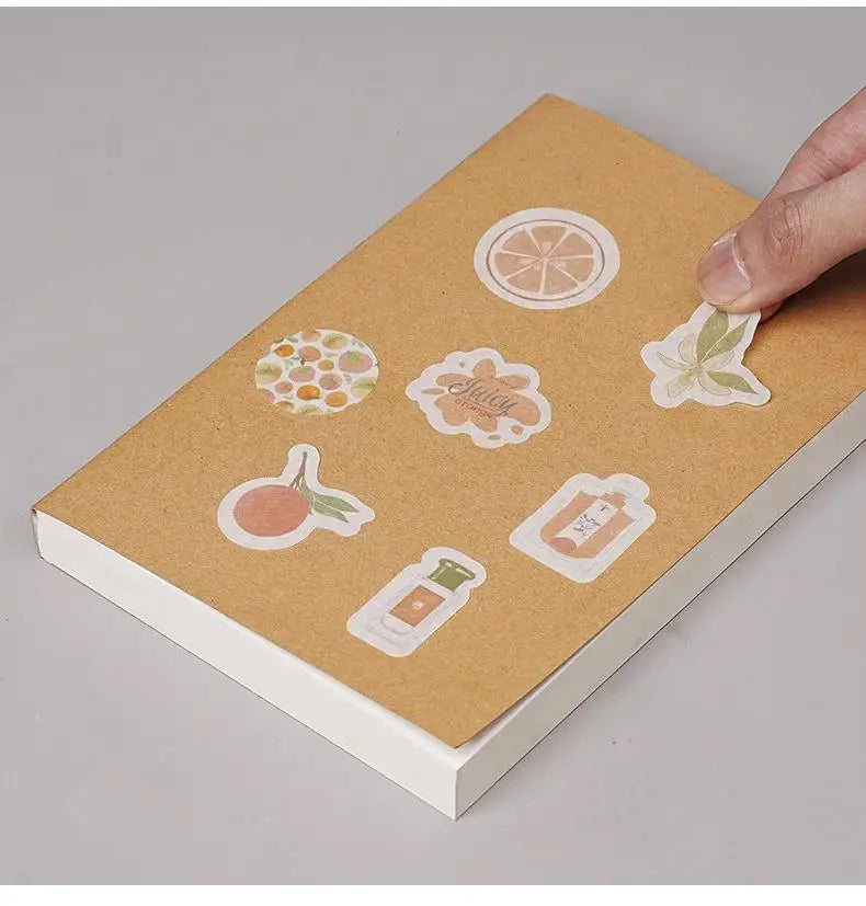 A hand places a finger on the cover of a kraft paper DIY notebook featuring various citrus and floral designs.