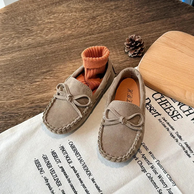 A pair of tan suede girls' moccasins with bow details resting on a white paper with wine tasting notes.