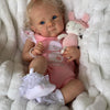 A baby doll with light hair and pink clothing is lying in white fluffy material next to a pink stuffed animal.