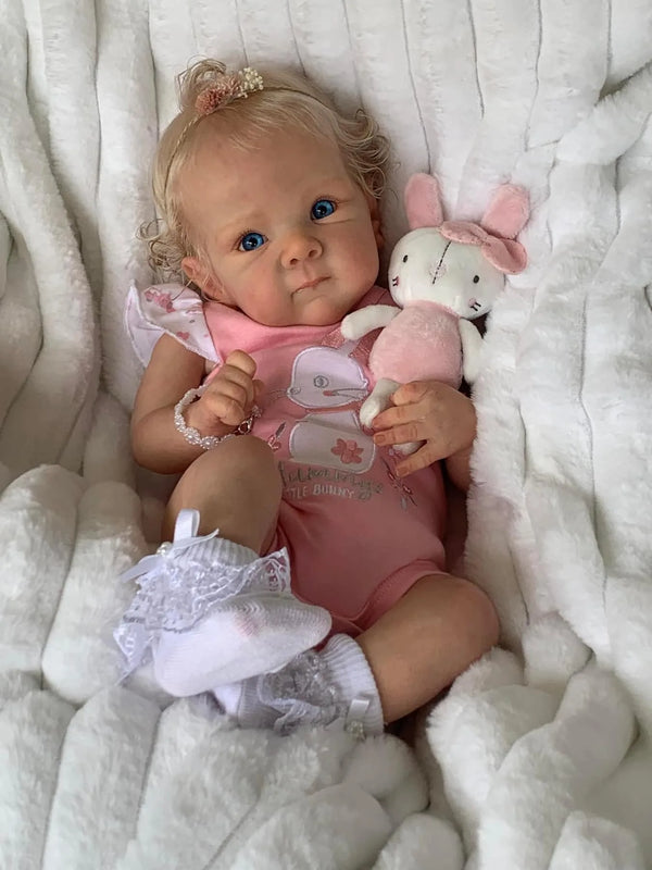 En babydokke med lyst hår og lyserødt tøj ligger i hvidt, fluffy materiale ved siden af et lyserødt bamse.