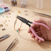 A hand holds black-handled jewelry pliers and cutters on a wooden surface with various beads and tools nearby.