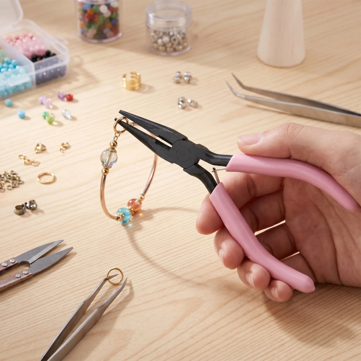 A hand holds black-handled jewelry pliers and cutters on a wooden surface with various beads and tools nearby.