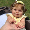 A close-up photo of a baby doll with light brown hair wearing a yellow top and a white flower headband.