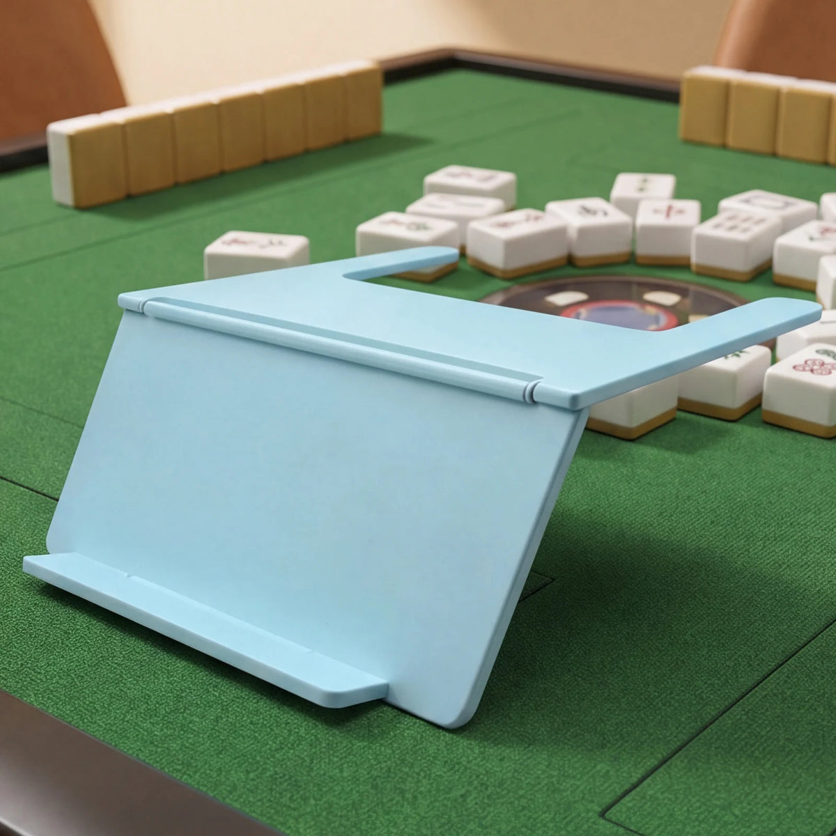 A light blue foldable mahjong card holder with a non-slip stand is placed on a green mat surrounded by white tiles.