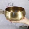 A hand holds a brass Tibetan singing bowl with hammered texture against a light background.