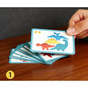 Une main tient une carte d'un jeu de dinosaures en bois, avec des dinosaures bleus et rouges, sur une surface en bois sombre.