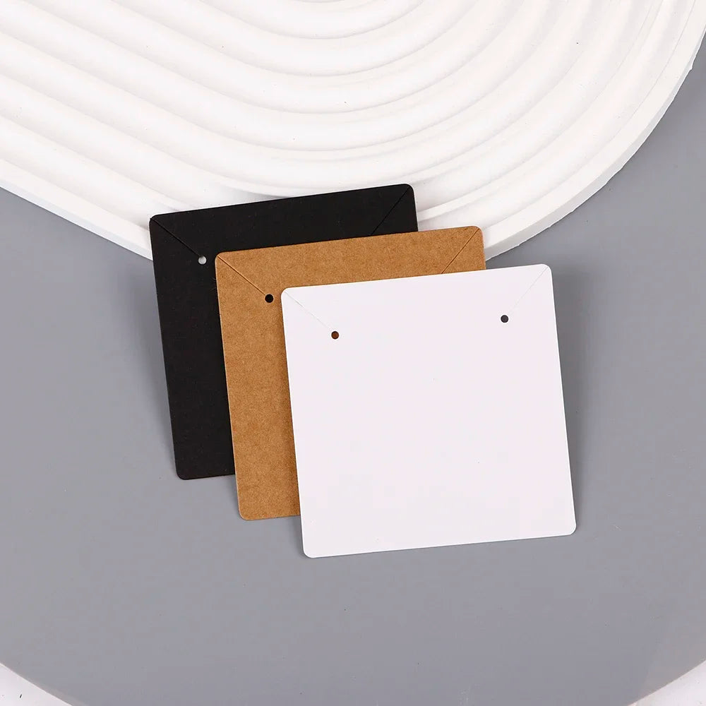Three square kraft jewelry display cards in black, brown, and white are displayed on a gray surface.