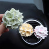 A hand holds a cluster of pale green silicone flower blossoms, with other light yellow and pink flowers visible in the backgr