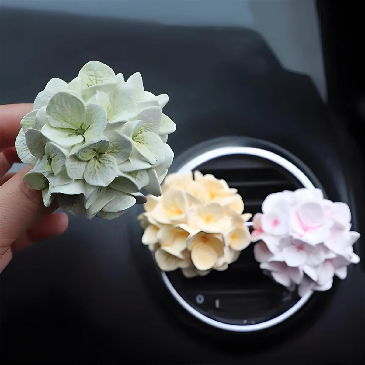 A hand holds a cluster of pale green silicone flower blossoms, with other light yellow and pink flowers visible in the backgr