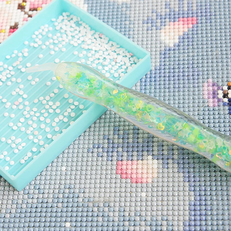 A glow-in-the-dark diamond painting pen with a translucent green body and colorful tips rests on a light blue tray.