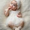 A newborn baby girl wearing a white lace tulle dress and hat, lying on a soft, light-colored surface.