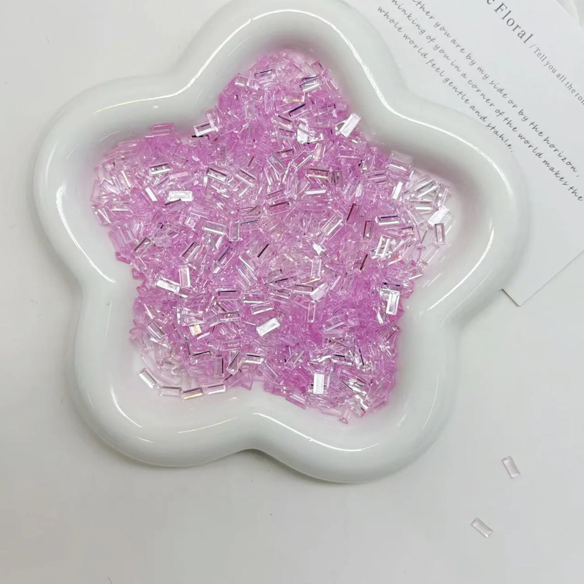 A white flower-shaped dish filled with pink glitter acrylic strips for nail art and DIY crafts