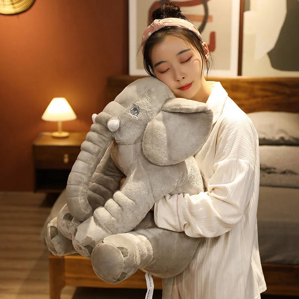 A woman is holding a large, gray plush elephant doll cushion in a warm indoor setting.