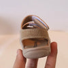 A hand holds a pair of tan soft-soled sandals for toddlers, showing the upper and sole.