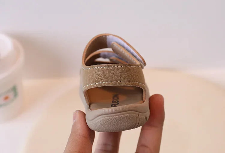 A hand holds a pair of tan soft-soled sandals for toddlers, showing the upper and sole.