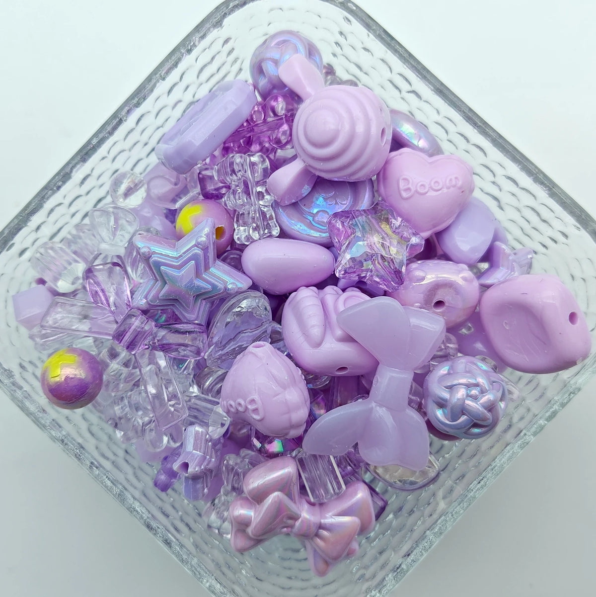 A clear glass container filled with various purple and pink acrylic loose beads for jewelry making