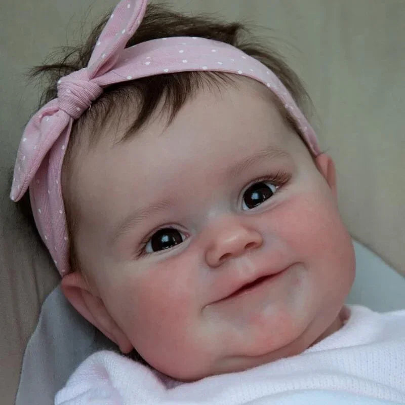 A close-up photo of a newborn reborn baby doll with pink hair tied in a bow, smiling.