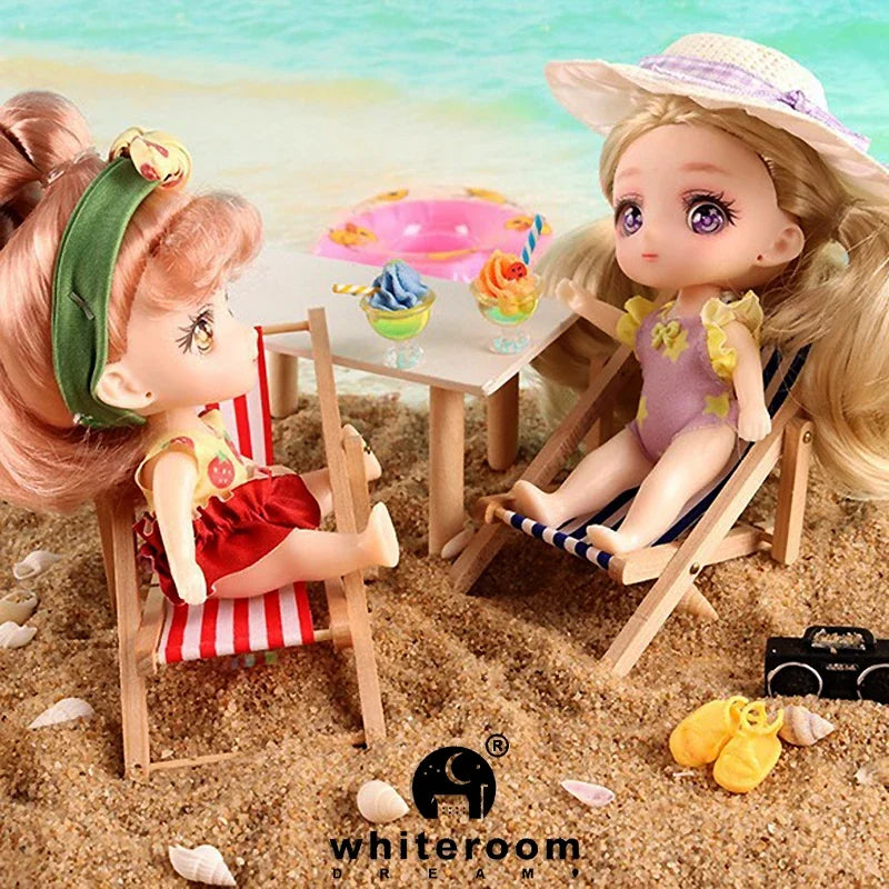 A miniature doll with pink hair and a floral hat is seated in a wooden chair on sand, next to a small pink ice cream cone.
