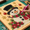 A hand is painting a detailed snowman design using red, green, and white pearls on a canvas board.