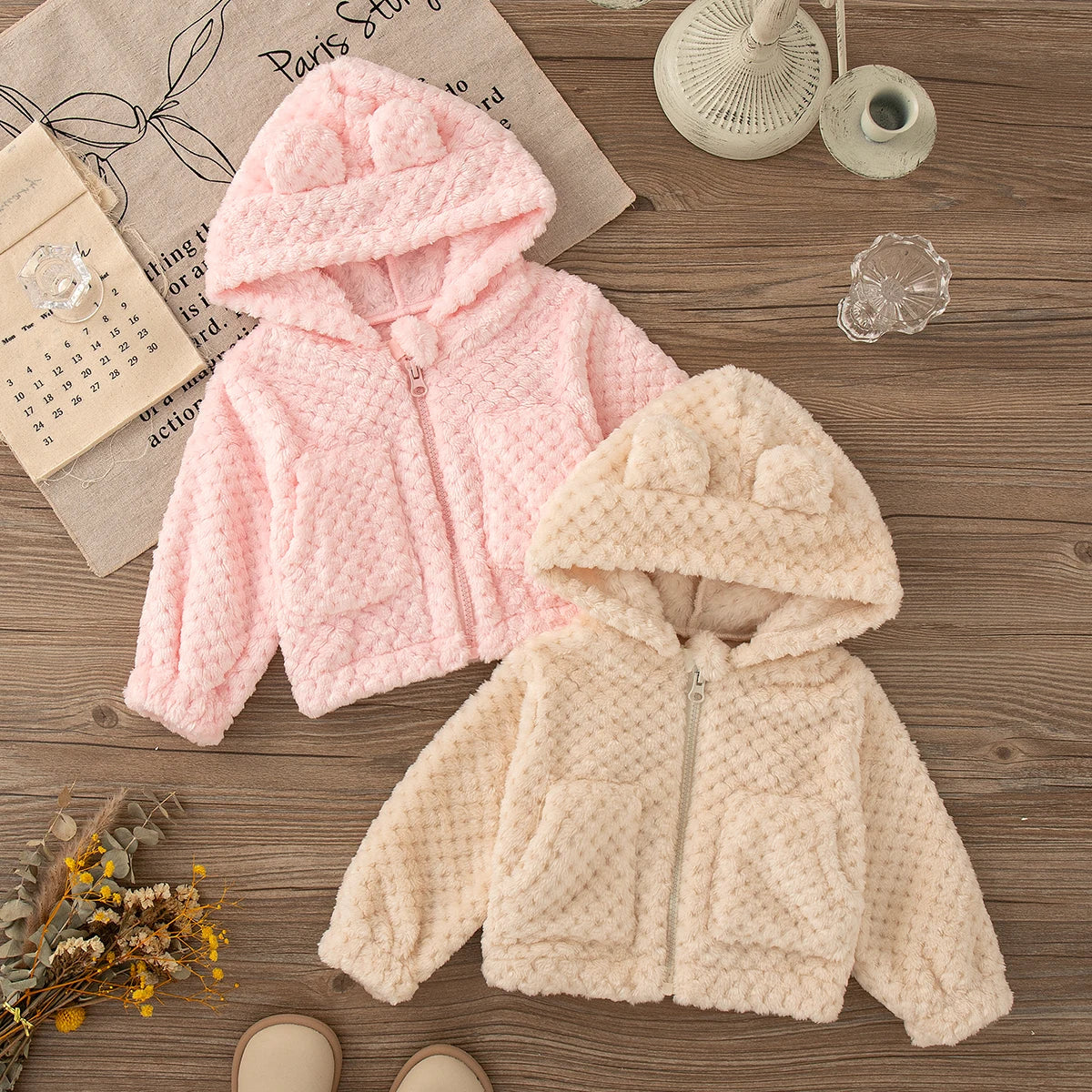 A pink and cream plush hooded winter jacket for a baby girl displayed on a wooden surface.