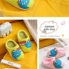 A set of yellow and blue cartoon dinosaur plush slippers for a baby, shown on a yellow fabric background.