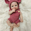 A realistic soft silicone reborn doll wearing a pink knitted bunny hat and outfit, lying on a white fluffy surface.