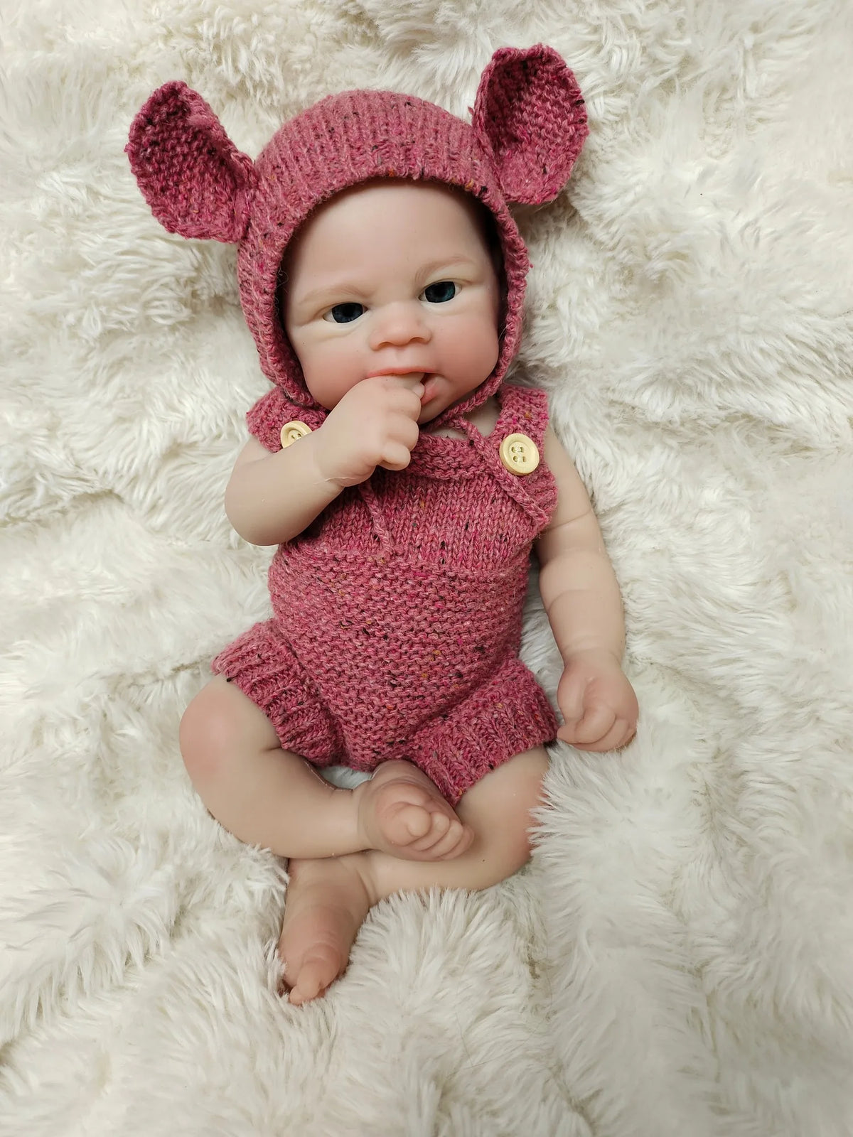 A realistic soft silicone reborn doll wearing a pink knitted bunny hat and outfit, lying on a white fluffy surface.
