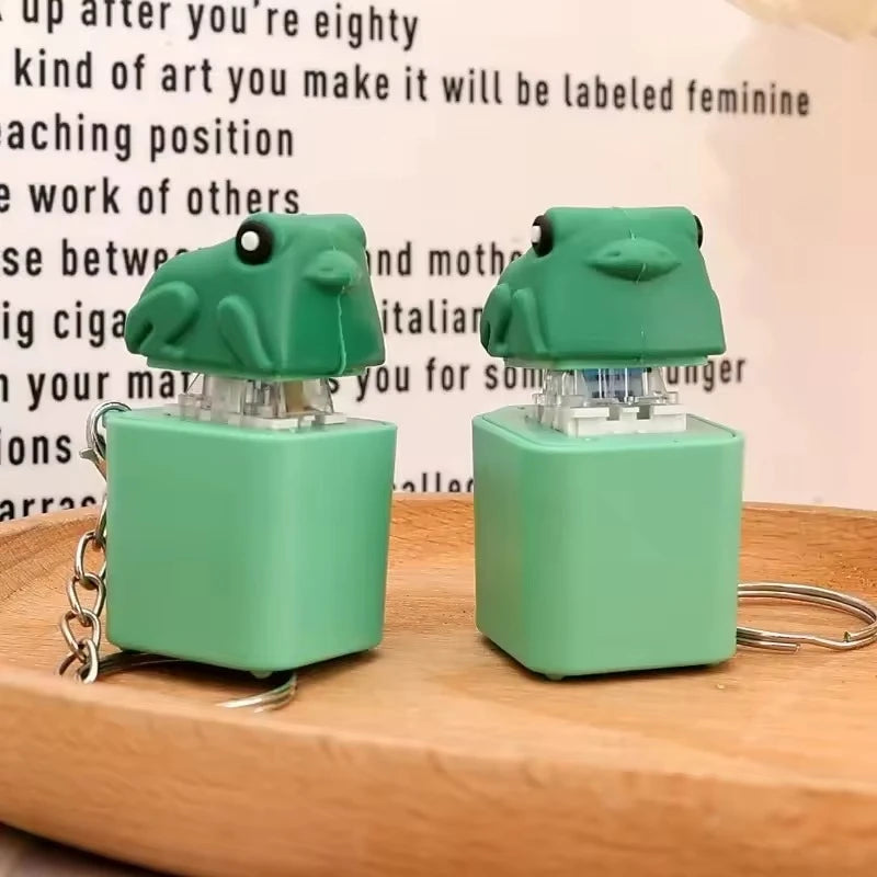 A pair of green duck and frog fidget clicker keychains resting on a wooden surface.