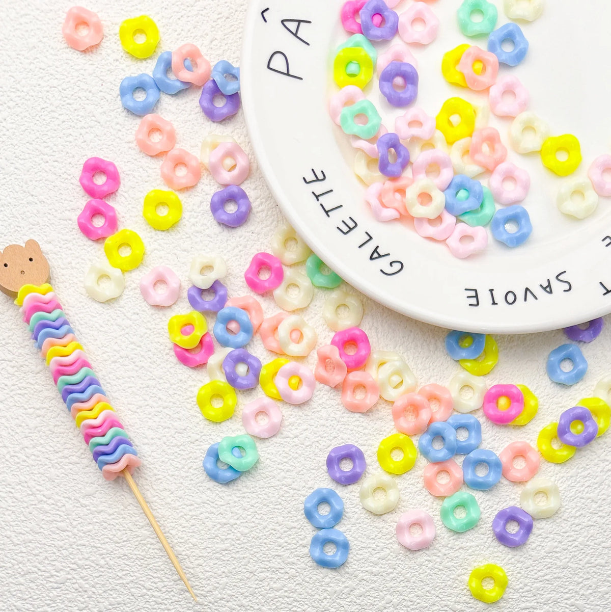 A collection of colorful pastel wavy acrylic beads in various shades are scattered on a white surface next to a white plate.