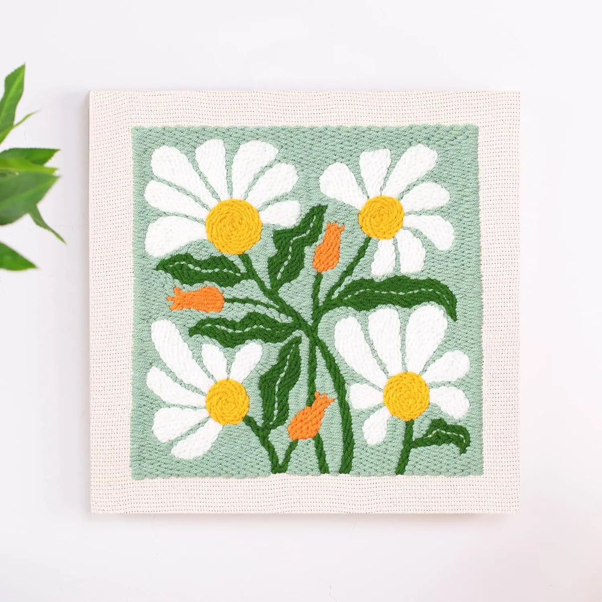 A square punch needle embroidery featuring white and yellow daisies on a pale green background.