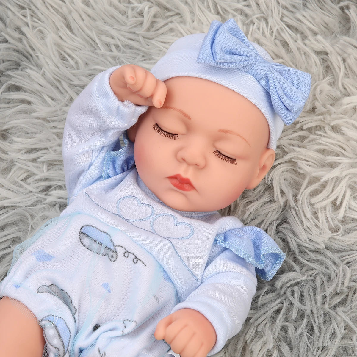 A lifelike soft vinyl reborn doll with a blue bow and matching outfit lying on a gray fuzzy surface.
