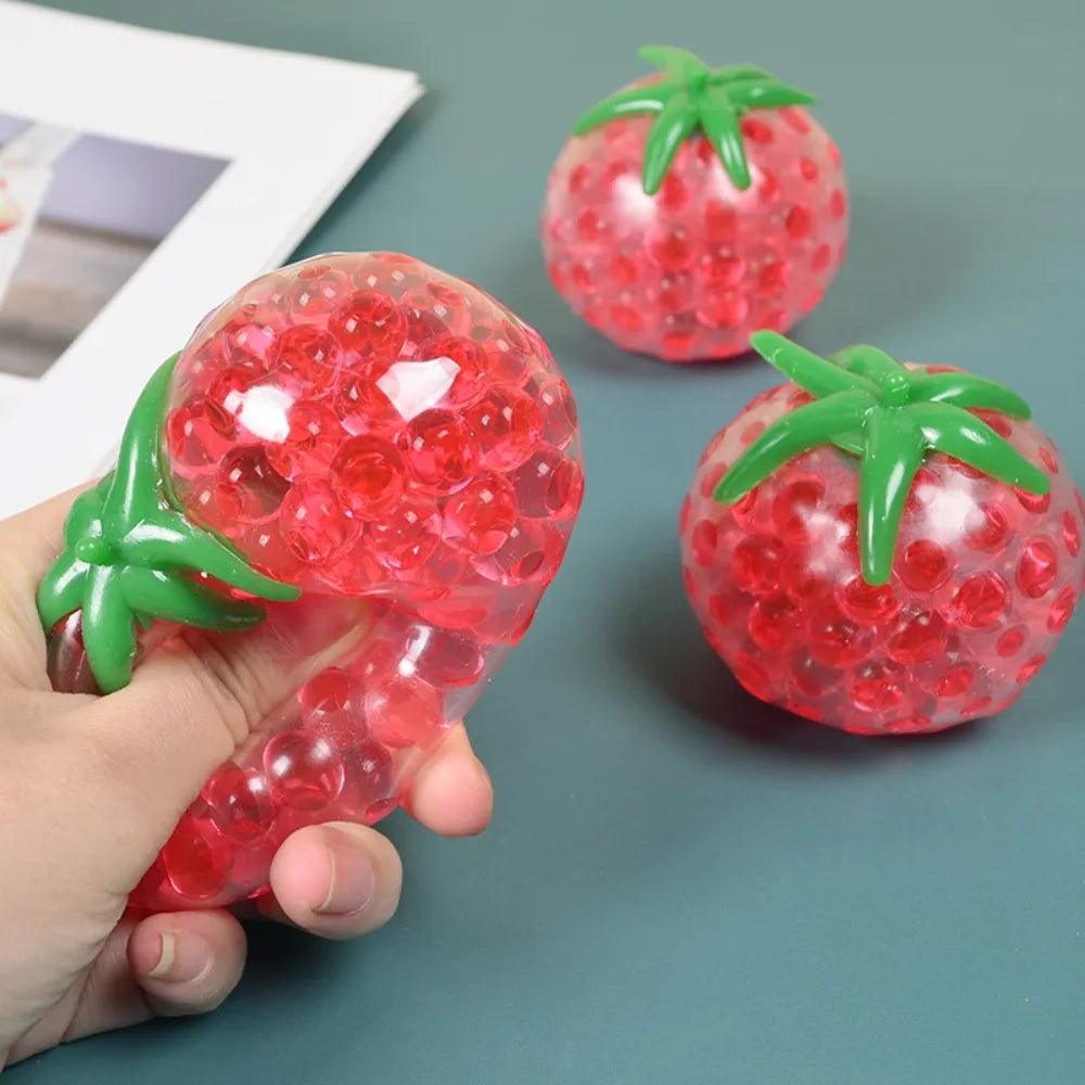 A hand holds a translucent red squishy tomato stress relief fidget toy with green leaves.
