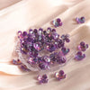 A pile of purple acrylic flower beads is displayed on a light fabric surface.