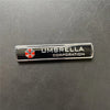 A black metal badge with the text UMBRELLA CORPORATION and a red and silver umbrella emblem.